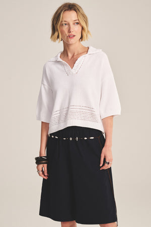 A woman stands against a plain background wearing the WINSLOW SWEATER POLO by Velvet by Graham & Spencer—a white cropped sweater polo with pointelle detailing—paired with a black skirt, shell belt, bracelets, and a ring.