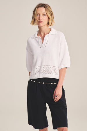 A woman wears the WINSLOW SWEATER POLO by Velvet by Graham & Spencer— a white cropped sweater polo with pointelle details—paired with black knee-length shorts, a thin belt, and rings, standing against a plain background.