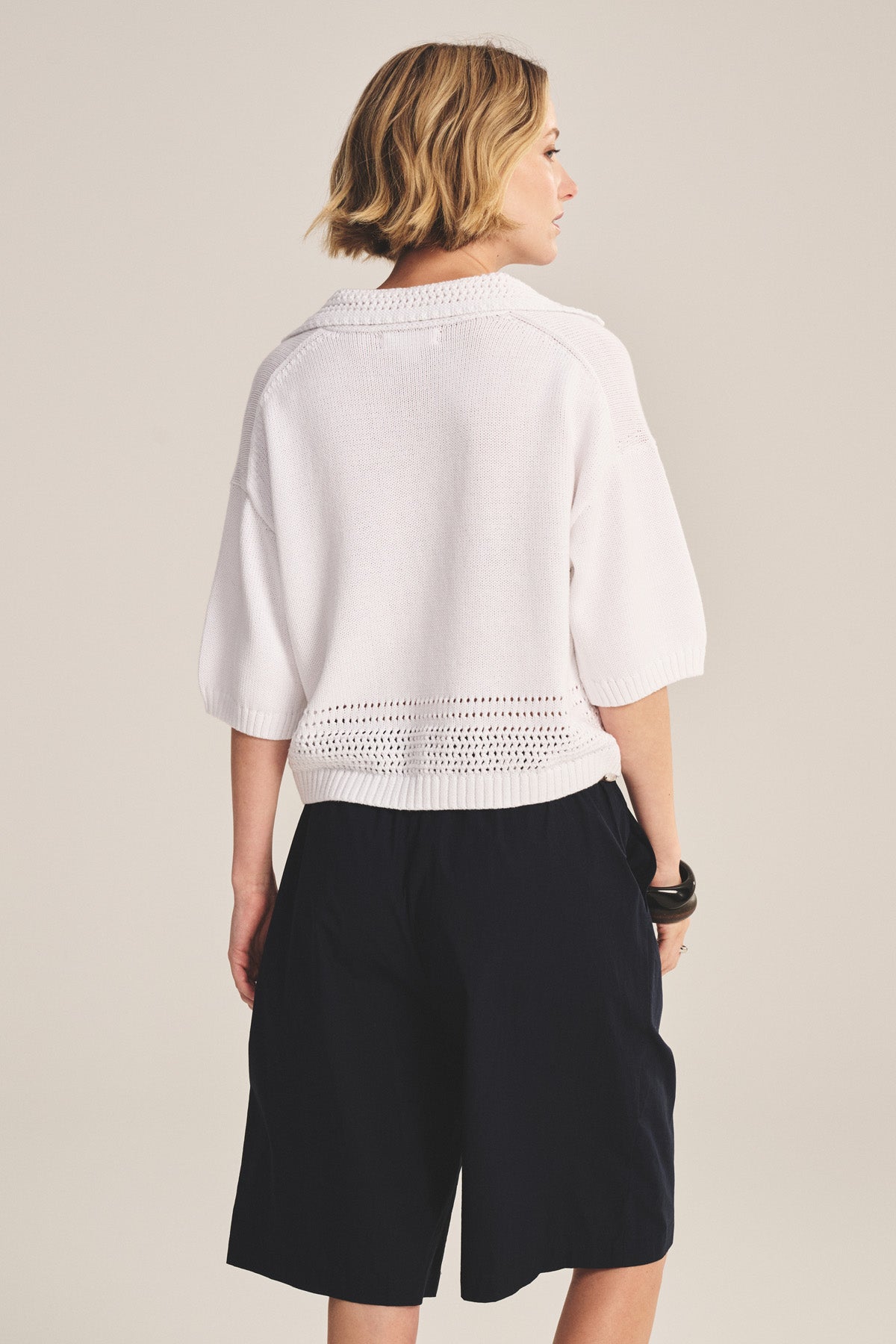 A person stands facing away, wearing the WINSLOW SWEATER POLO by Velvet by Graham & Spencer in white pima cotton with pointelle details, paired with loose black knee-length shorts against a plain background.