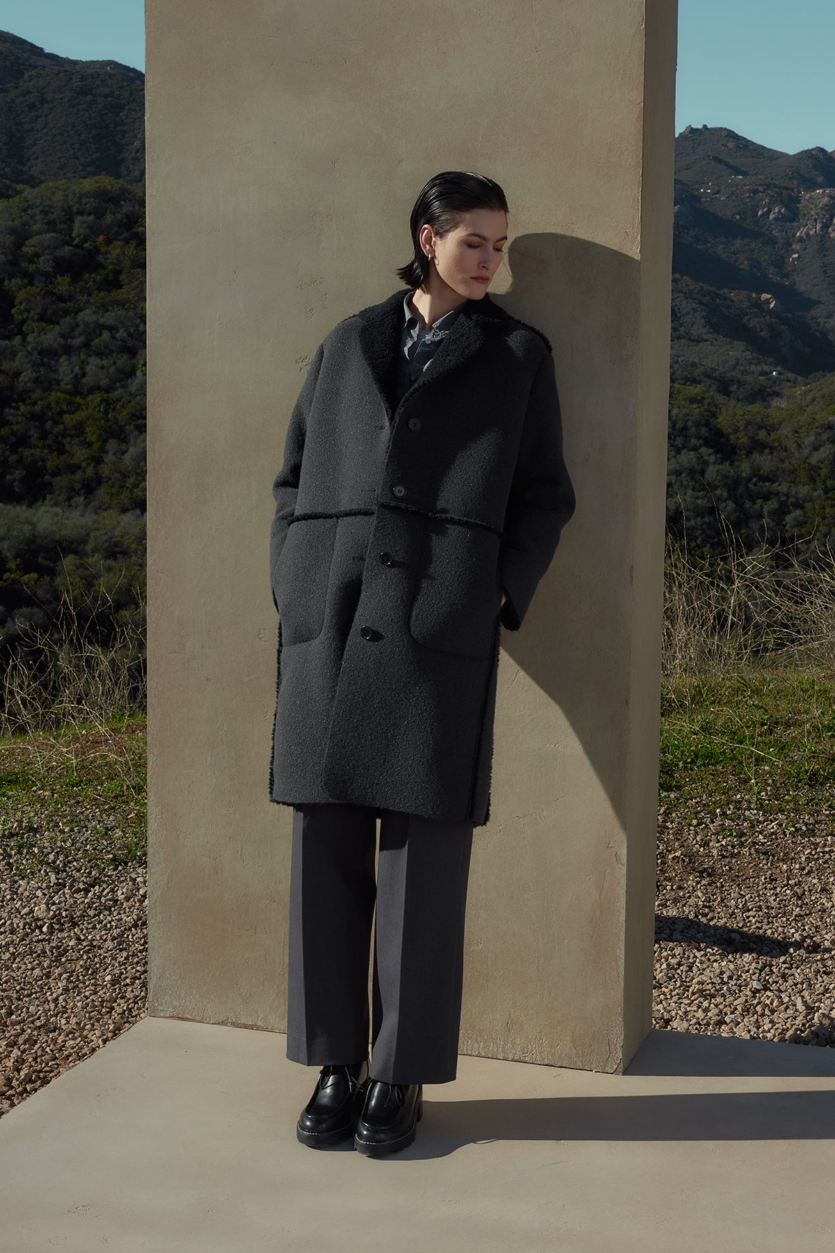   A person with short dark hair wearing the ROSEMEAD COAT by Velvet by Jenny Graham stands against a stone wall in an outdoor setting with mountains in the background. 