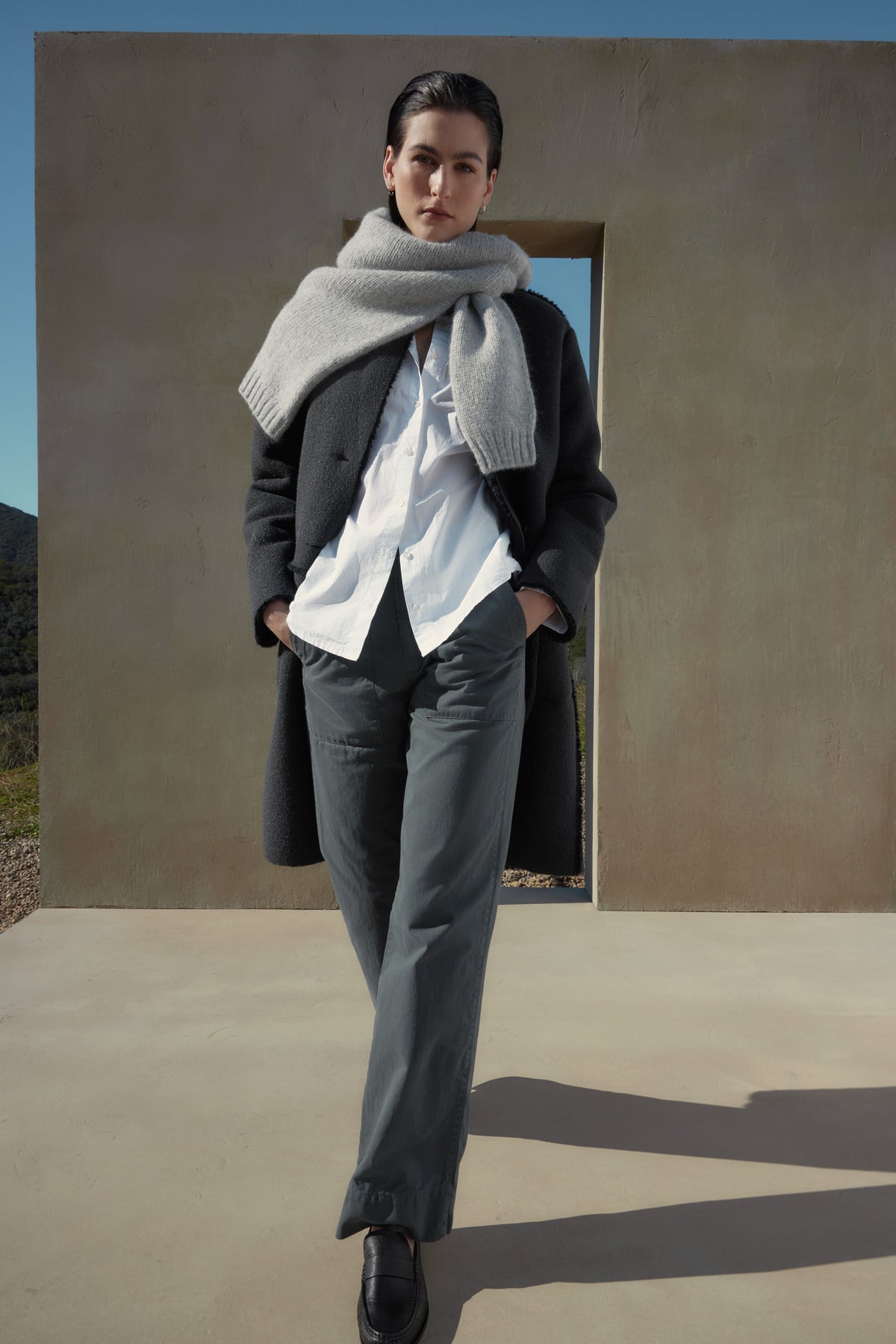 A person stands outdoors, wearing a dark coat, white shirt, cotton twill scarf, and the VENTURA PANT by Velvet by Jenny Graham. A concrete structure with a rectangular opening is behind them.-37665534804161