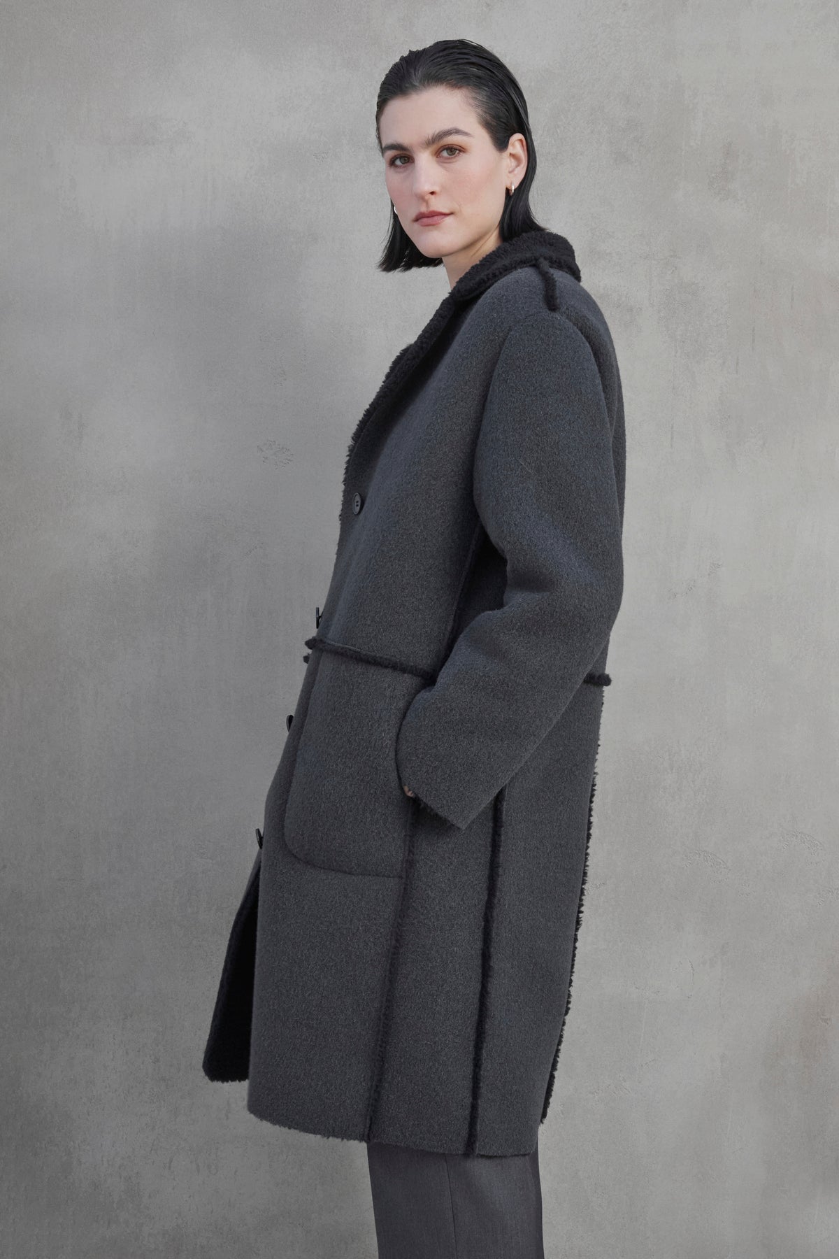   A person with dark hair stands against a gray backdrop, wearing the ROSEMEAD COAT, a long, dark gray wool coat by Velvet by Jenny Graham, featuring large front pockets and black trim, looking slightly to the side. 