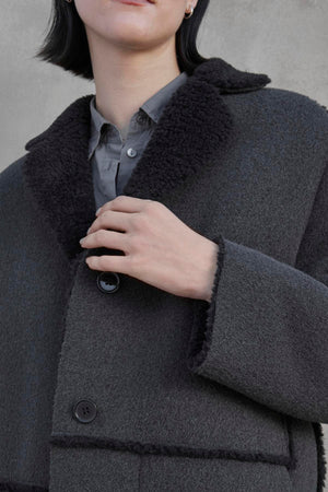 An individual with short dark hair is wearing the ROSEMEAD COAT by Velvet by Jenny Graham, which features large buttons and a black shearling collar, over a grey collared shirt.