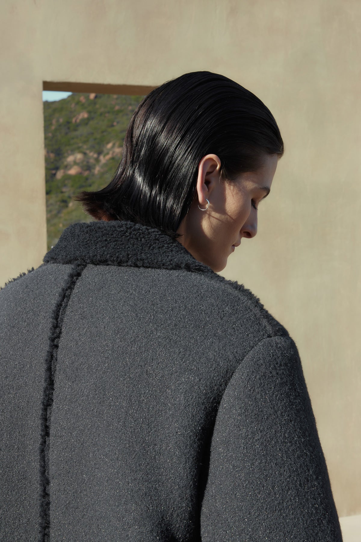   A person with dark, slicked-back hair is seen from behind, wearing the ROSEMEAD COAT by Velvet by Jenny Graham. The background features an outdoor setting with subtle landscape elements visible through an opening. 