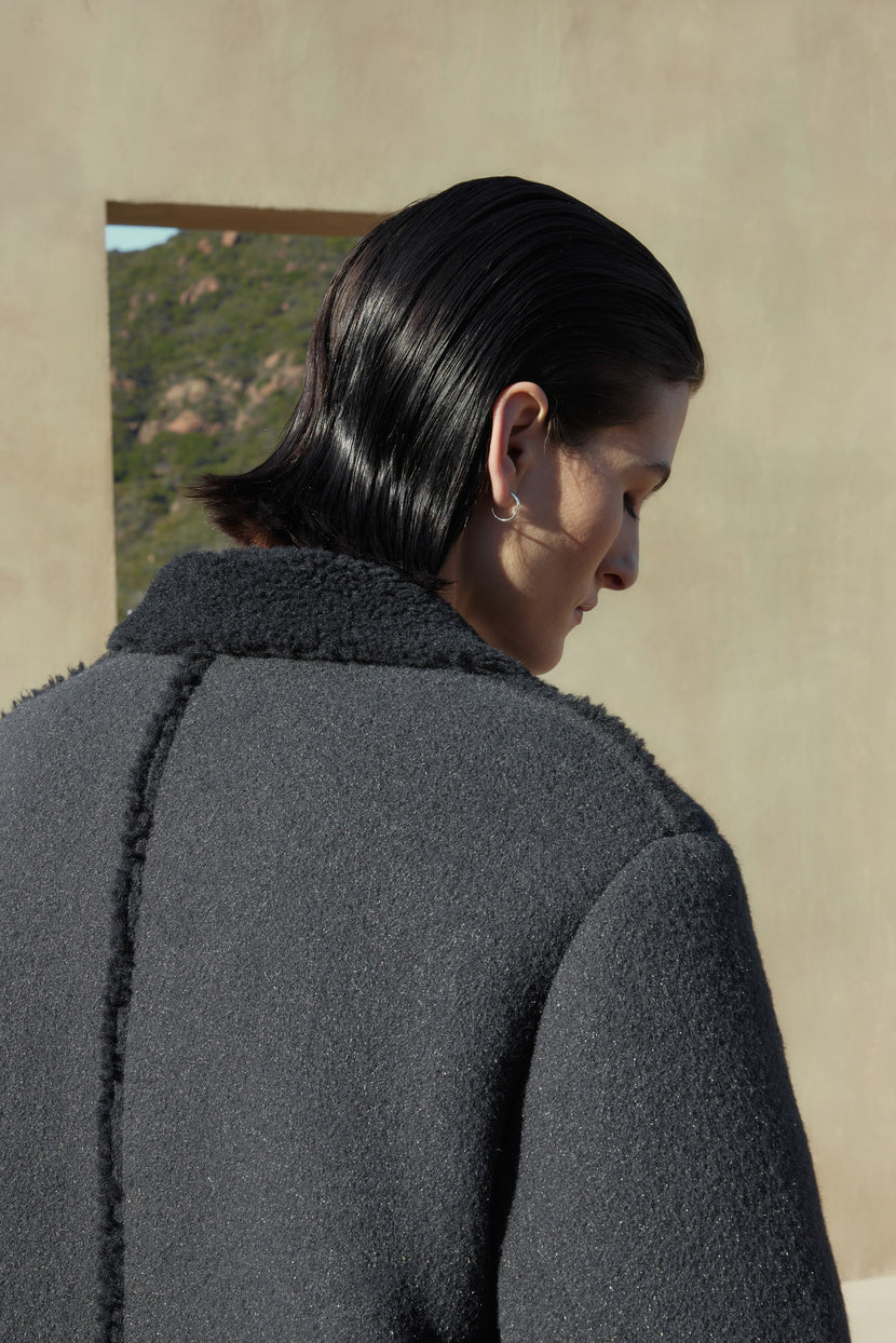 A person with dark, slicked-back hair is seen from behind, wearing the ROSEMEAD COAT by Velvet by Jenny Graham. The background features an outdoor setting with subtle landscape elements visible through an opening.