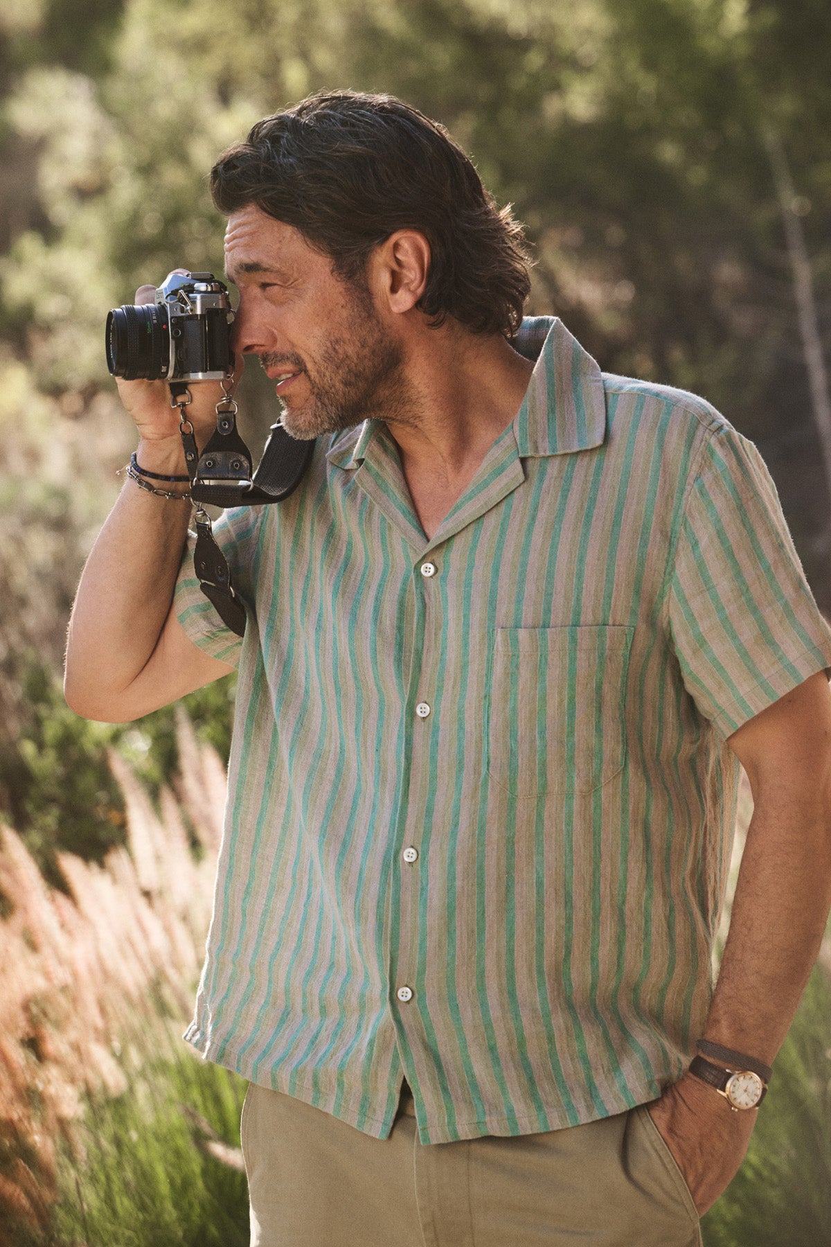 A man outdoors in sunlight and greenery holds a camera, wearing the WORTH SHIRT by Velvet by Graham & Spencer—a green and beige striped camp-collar shirt.-41297081336001