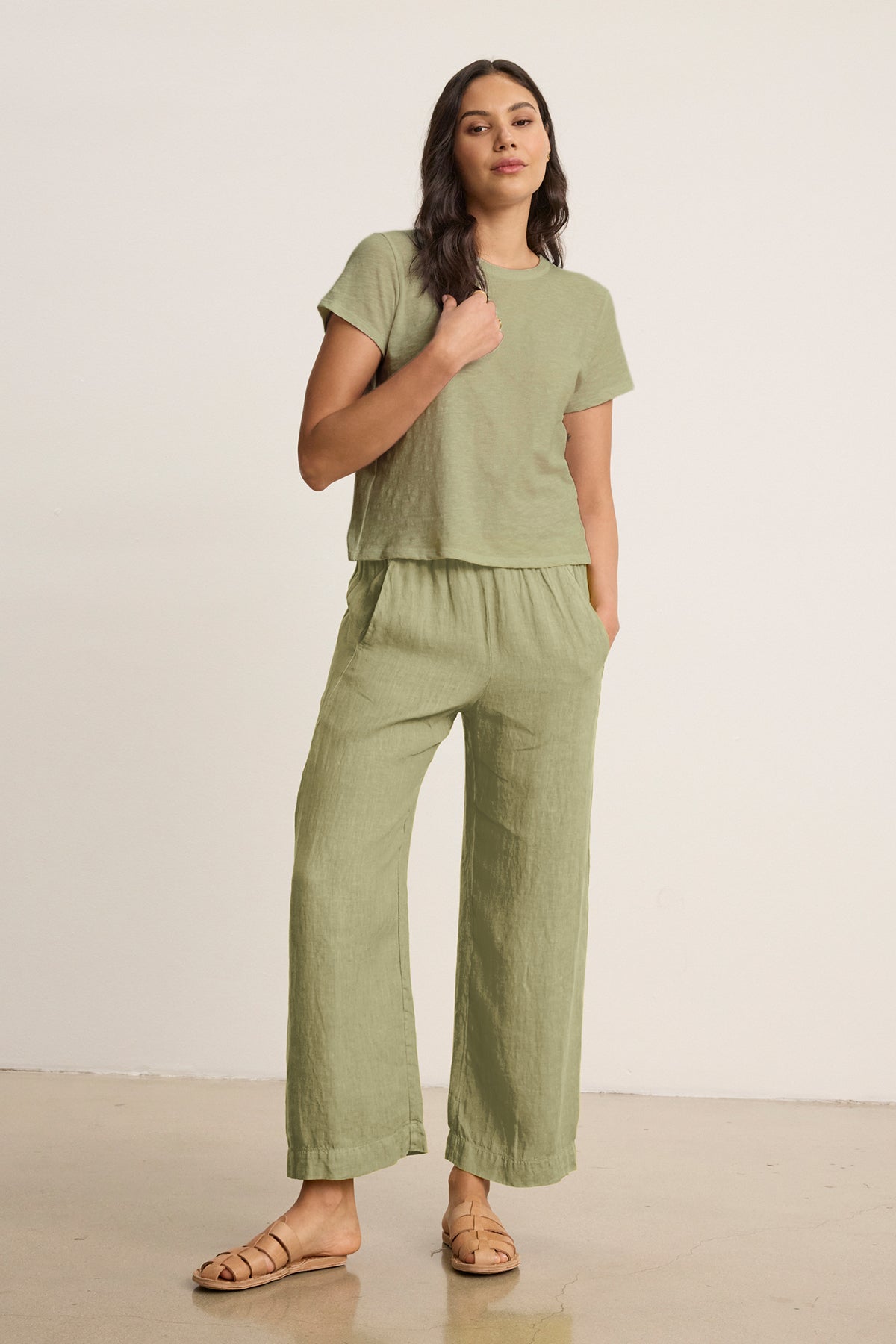 A woman wears the Velvet by Graham & Spencer LOLA LINEN PANT—lightweight, pull-on linen trousers with an elastic waist—in light green, paired with a matching short-sleeve top and sandals against a plain background.-39359712952513