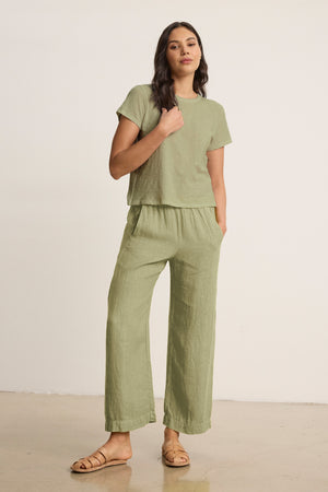 A woman wears the Velvet by Graham & Spencer LOLA LINEN PANT—lightweight, pull-on linen trousers with an elastic waist—in light green, paired with a matching short-sleeve top and sandals against a plain background.