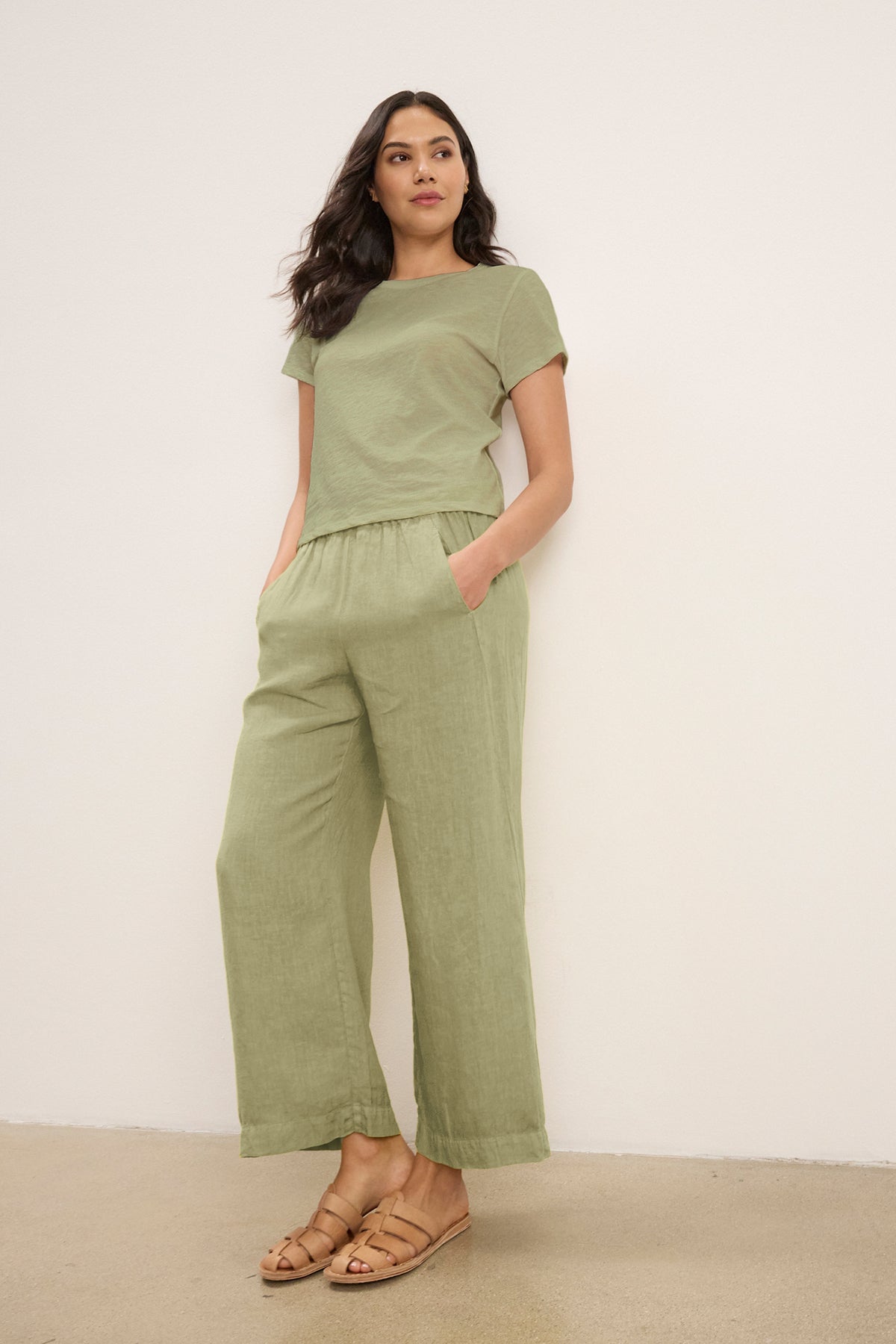 A woman stands against a plain wall wearing Velvet by Graham & Spencer's LOLA LINEN PANT in light green, paired with a matching short-sleeve top and tan sandals, her hands tucked in the pants' pockets.-39359713018049