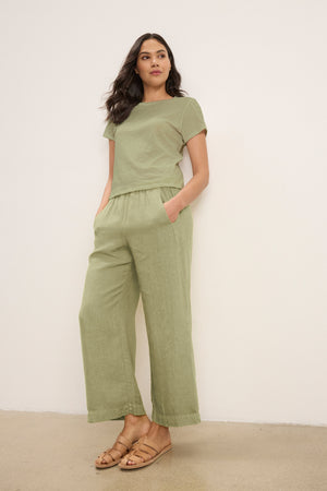 A woman stands against a plain wall wearing Velvet by Graham & Spencer's LOLA LINEN PANT in light green, paired with a matching short-sleeve top and tan sandals, her hands tucked in the pants' pockets.