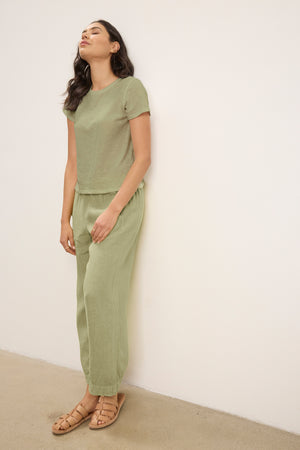 A woman stands against a plain white wall wearing Velvet by Graham & Spencer's LOLA LINEN PANT in light green, paired with a matching short-sleeve linen top and tan sandals.