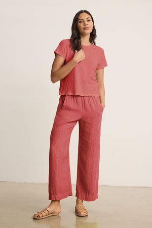 A woman stands against a plain wall wearing the Velvet by Graham & Spencer LOLA LINEN PANT, a lightweight, elastic-waist pull-on trouser paired with a matching pink short-sleeve top and tan sandals.