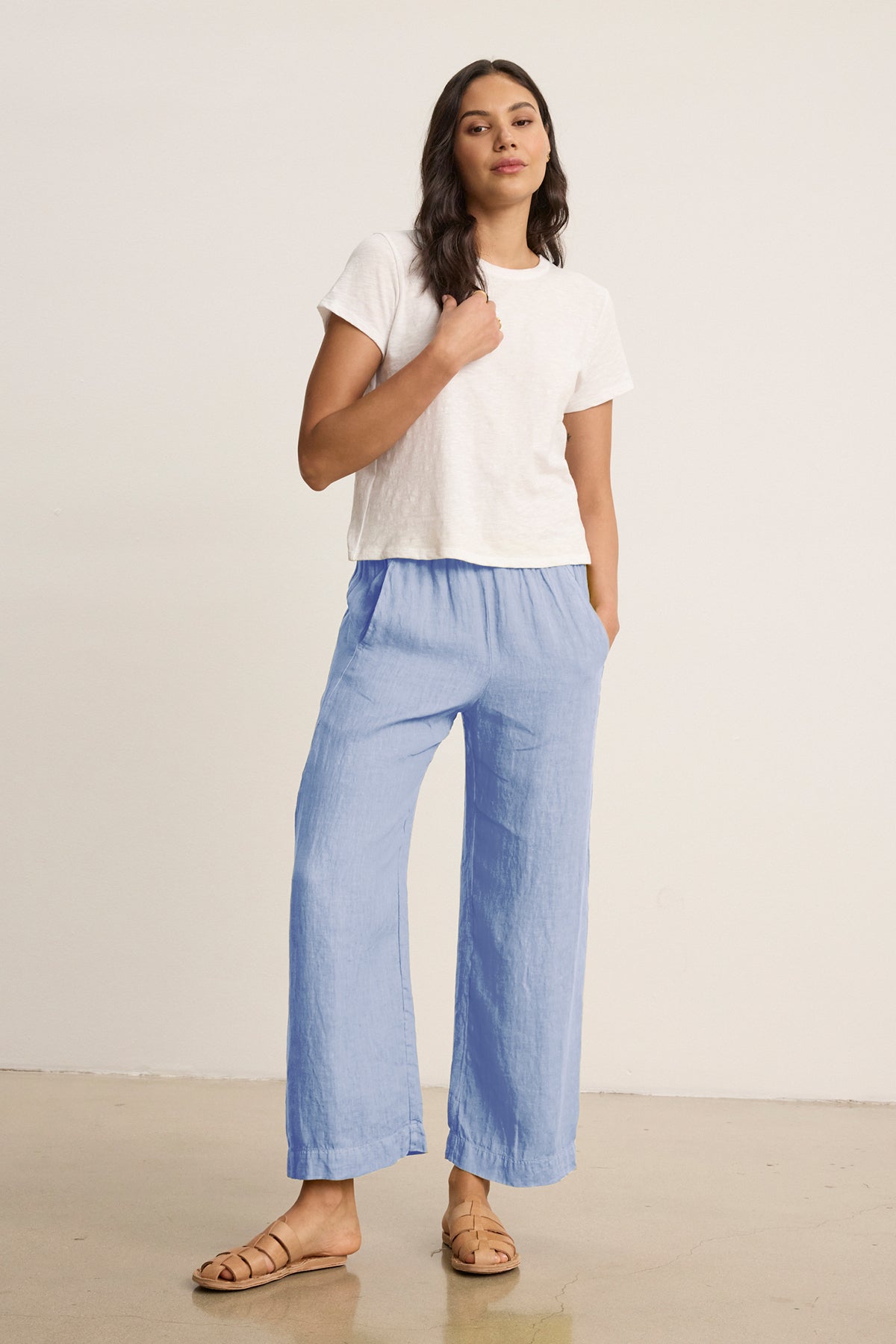 A woman stands indoors wearing a white short-sleeve t-shirt, tan sandals, and Velvet by Graham & Spencer’s LOLA LINEN PANT in light blue with an elastic waist, with one hand in her pocket.-39333424693441