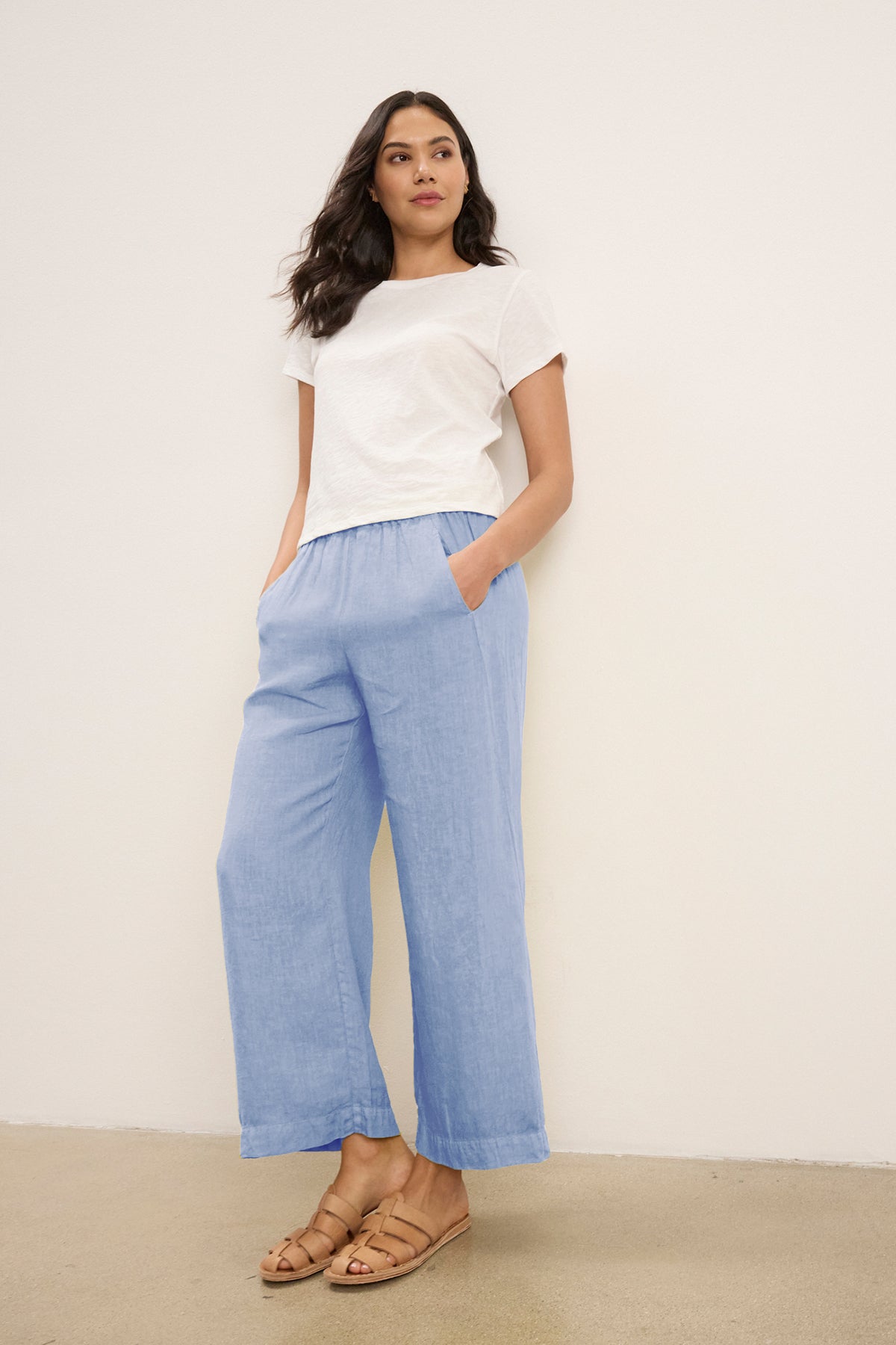   A woman stands against a white wall wearing a white t-shirt, tan sandals, and Velvet by Graham & Spencer's LOLA LINEN PANT in light blue with an elastic waist, hands casually in her pockets. 