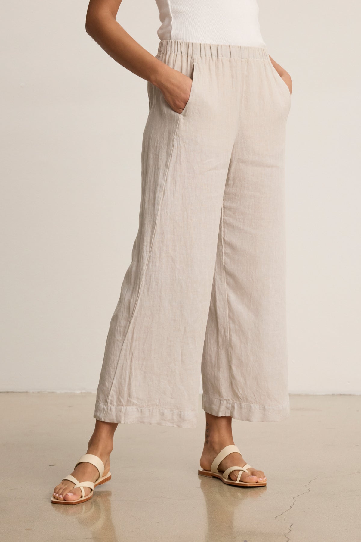 Wearing Velvet by Graham & Spencer’s LOLA LINEN PANT, a person stands on a smooth floor with hands in pockets, styled with a white sleeveless top and beige sandals.-39333428101313