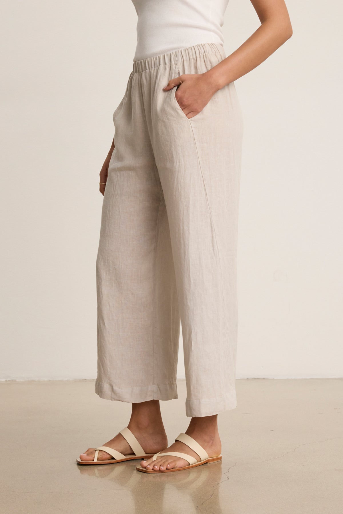 Wearing the Velvet by Graham & Spencer LOLA LINEN PANT, a person with a white sleeveless top and beige sandals stands indoors, one hand in a pocket.-39333428068545