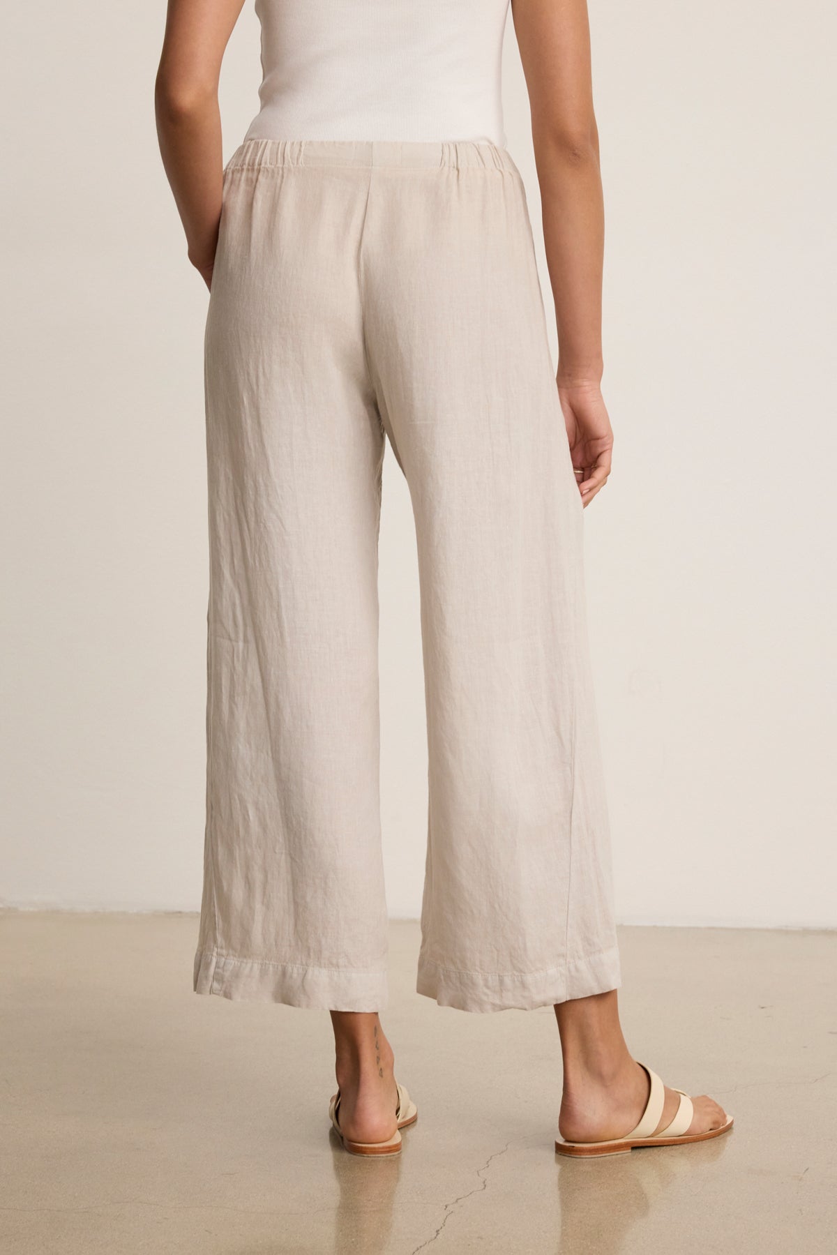 A person in Velvet by Graham & Spencer’s LOLA LINEN PANT, a white sleeveless top, and tan slide sandals stands on a polished floor facing away from the camera.-39333428035777