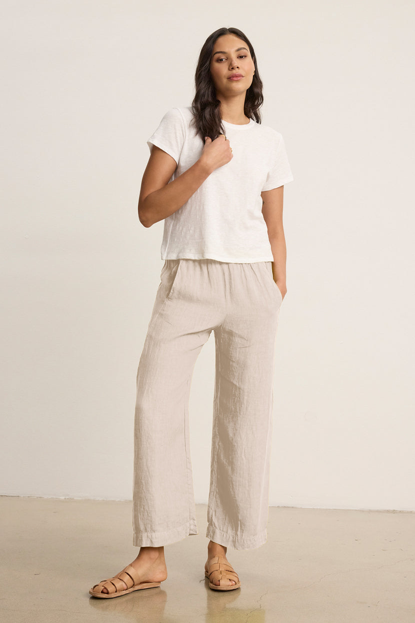 A woman stands indoors against a plain background, wearing a white short-sleeve t-shirt, tan sandals, and the LOLA LINEN PANT by Velvet by Graham & Spencer—lightweight pull-on trousers with an elastic waist.