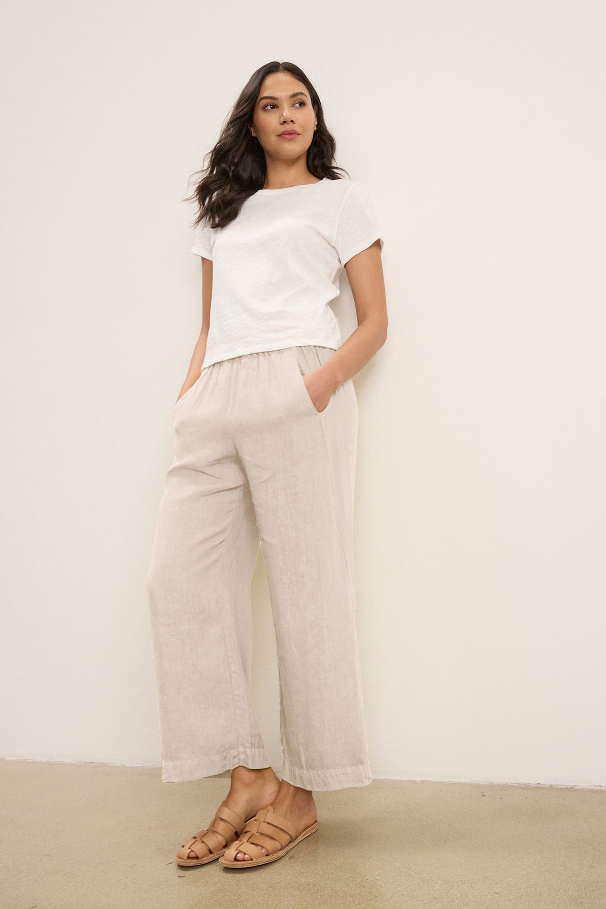 A woman stands against a plain wall, hands in pockets, wearing a white tee, tan sandals, and the LOLA LINEN PANT by Velvet by Graham & Spencer—lightweight pull-on trousers with an elastic waist.-39333427904705