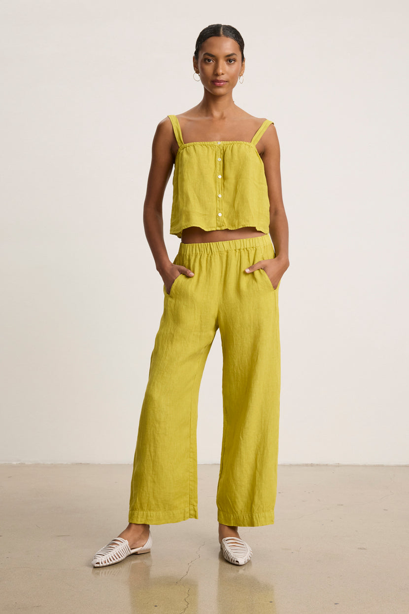A woman stands against a plain background wearing the LOLA LINEN PANT by Velvet by Graham & Spencer, a yellow sleeveless buttoned crop top, and white woven shoes.