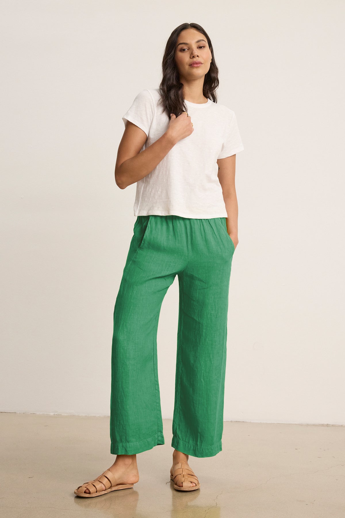 A woman stands indoors against a plain background wearing a white short-sleeve tee, tan sandals, and the LOLA LINEN PANT by Velvet by Graham & Spencer—green pull-on trousers with an elastic waist, her hands in her pockets.-39333752406209
