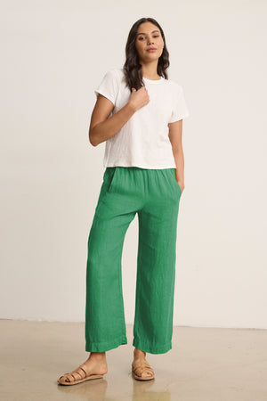 A woman stands indoors against a plain background wearing a white short-sleeve tee, tan sandals, and the LOLA LINEN PANT by Velvet by Graham & Spencer—green pull-on trousers with an elastic waist, her hands in her pockets.
