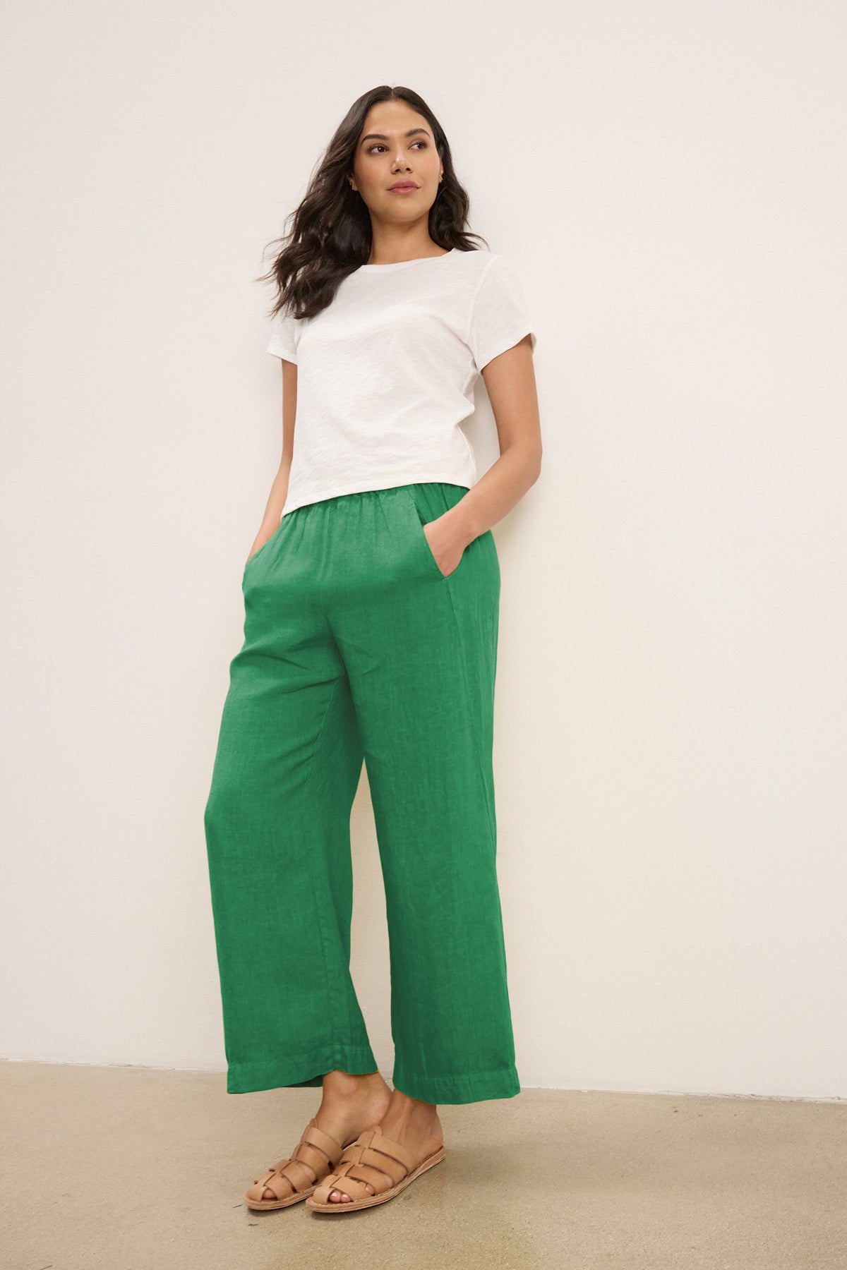 A person stands against a plain white wall with hands in pockets, wearing a white short-sleeve t-shirt, tan sandals, and the LOLA LINEN PANT by Velvet by Graham & Spencer.-39333752373441