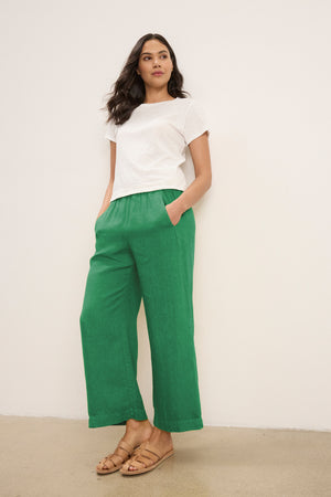 A person stands against a plain white wall with hands in pockets, wearing a white short-sleeve t-shirt, tan sandals, and the LOLA LINEN PANT by Velvet by Graham & Spencer.