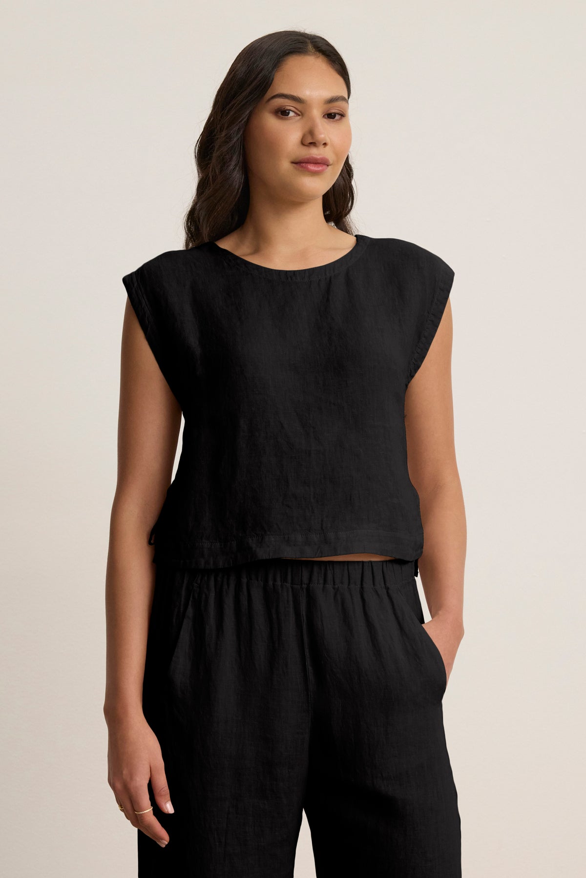 A woman stands against a plain background wearing the Velvet by Graham & Spencer TAVIA LINEN TOP, a sleeveless black cropped linen top, paired with matching black linen pants, one hand in her pocket.-39148767707329