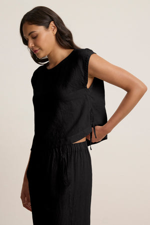 A woman wearing the TAVIA LINEN TOP by Velvet by Graham & Spencer—a black, sleeveless, cropped linen top with a matching skirt—stands with one hand adjusting the back of her top while looking down.