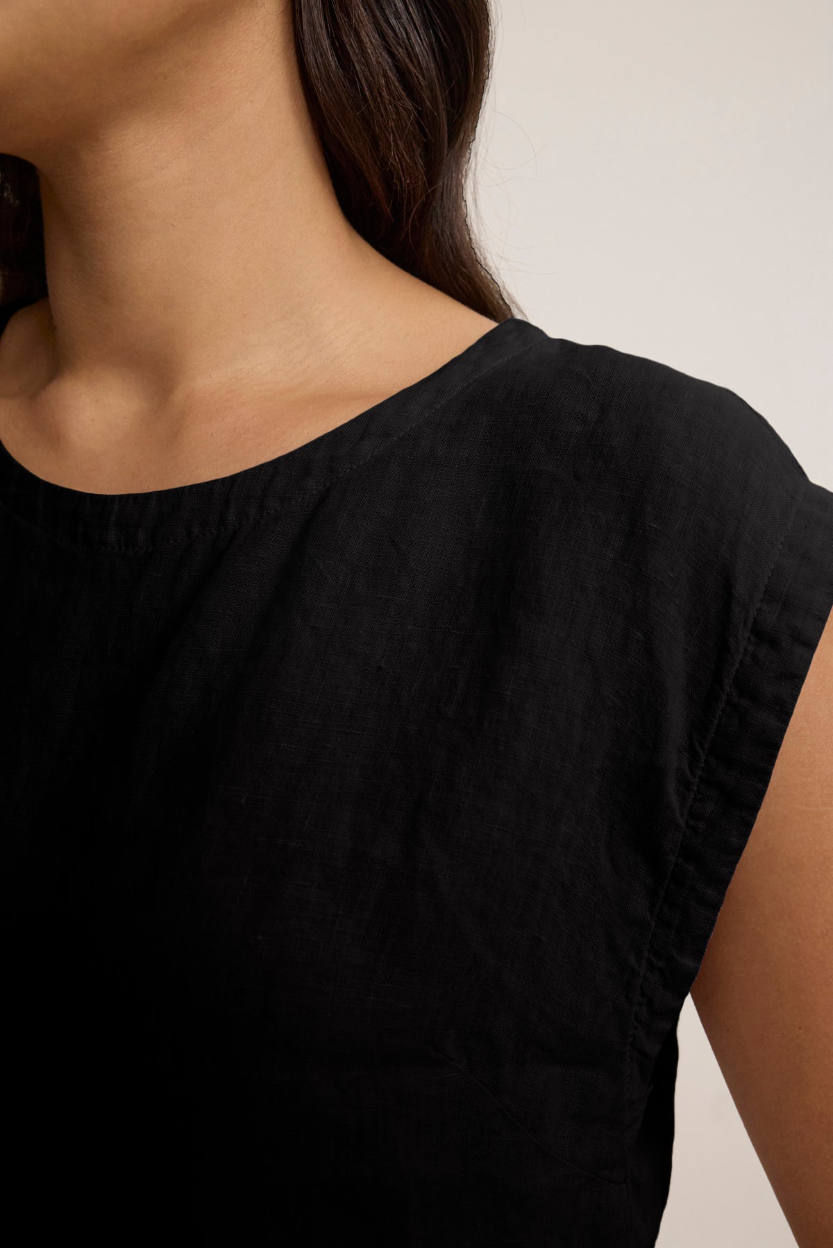 Close-up of a person wearing the TAVIA LINEN TOP by Velvet by Graham & Spencer—a black, short-sleeved, crew-neck linen top with a cropped fit—showing the upper torso and part of the neck and hair against a neutral background.-39148767477953