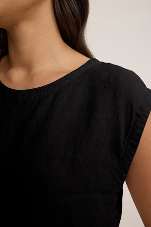Close-up of a person wearing the TAVIA LINEN TOP by Velvet by Graham & Spencer—a black, short-sleeved, crew-neck linen top with a cropped fit—showing the upper torso and part of the neck and hair against a neutral background.