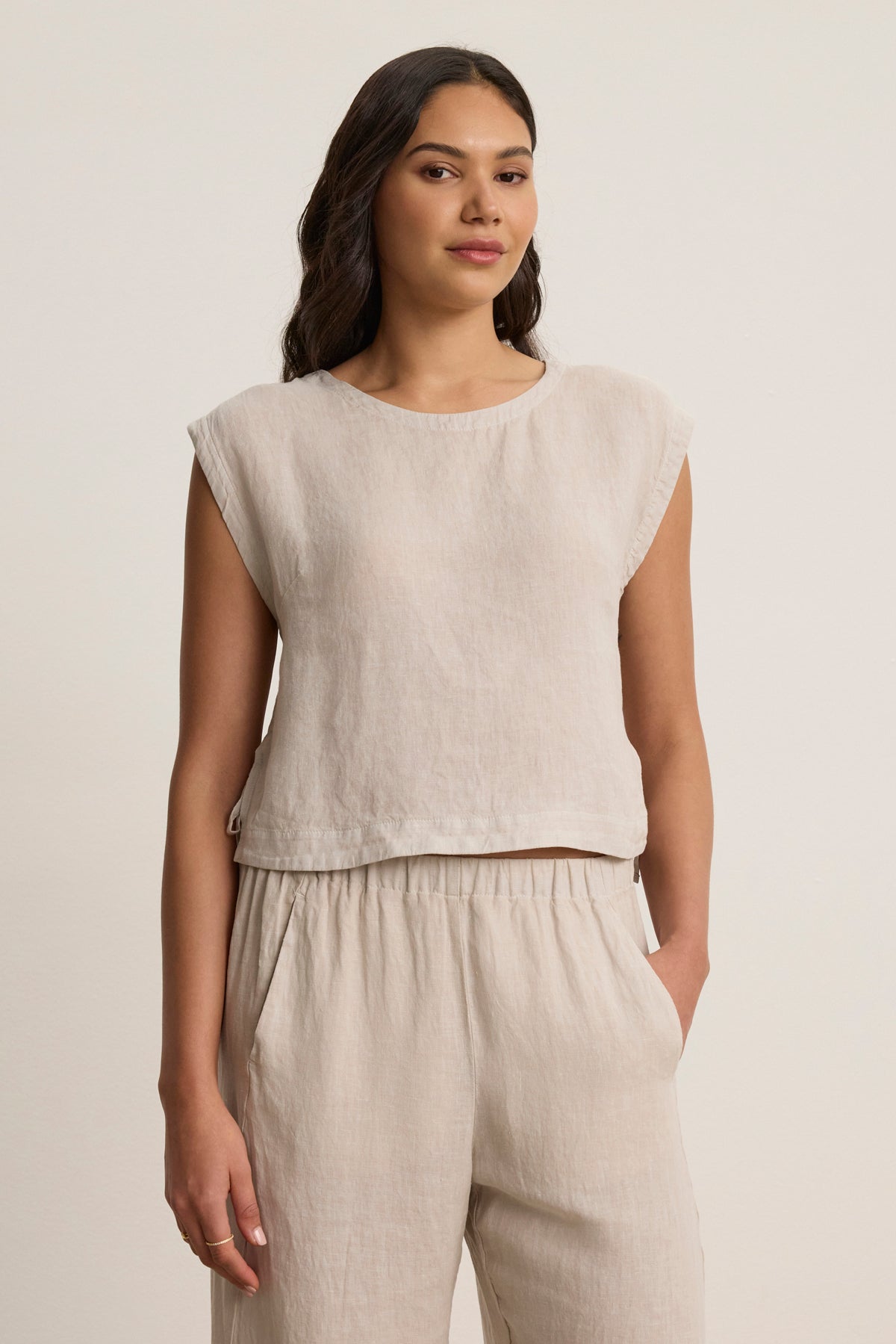 A woman stands against a plain background wearing the TAVIA LINEN TOP by Velvet by Graham & Spencer—a sleeveless, light beige cropped fit linen top that's stylish, breathable, and perfect for warm days.-39148767740097