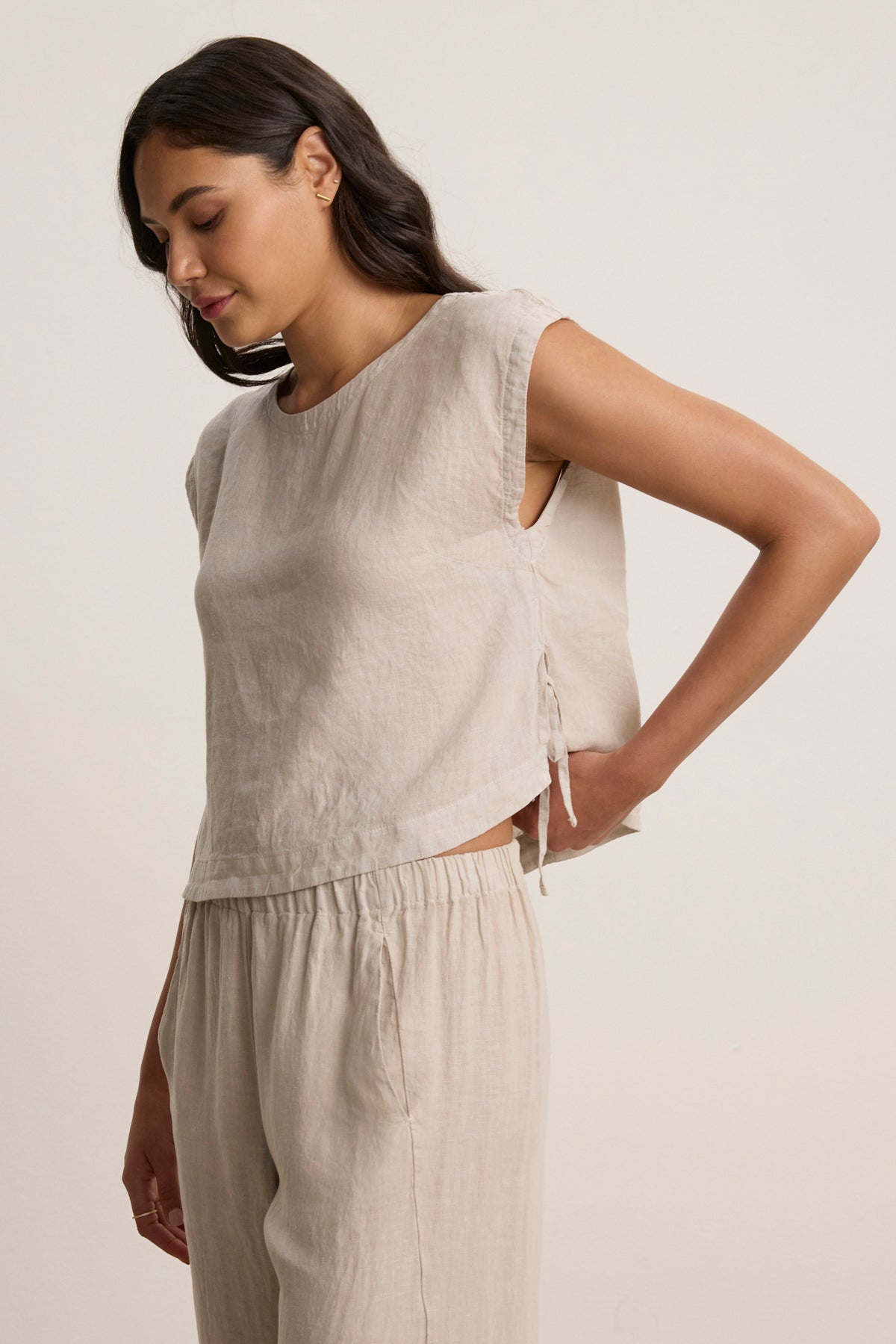 A woman wears the TAVIA LINEN TOP by Velvet by Graham & Spencer—a sleeveless, breathable, cropped linen top—paired with matching pants, standing against a plain background and looking downward with one hand on her hip.-39148767674561