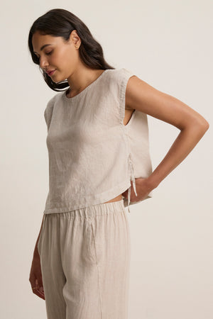 A woman wears the TAVIA LINEN TOP by Velvet by Graham & Spencer—a sleeveless, breathable, cropped linen top—paired with matching pants, standing against a plain background and looking downward with one hand on her hip.