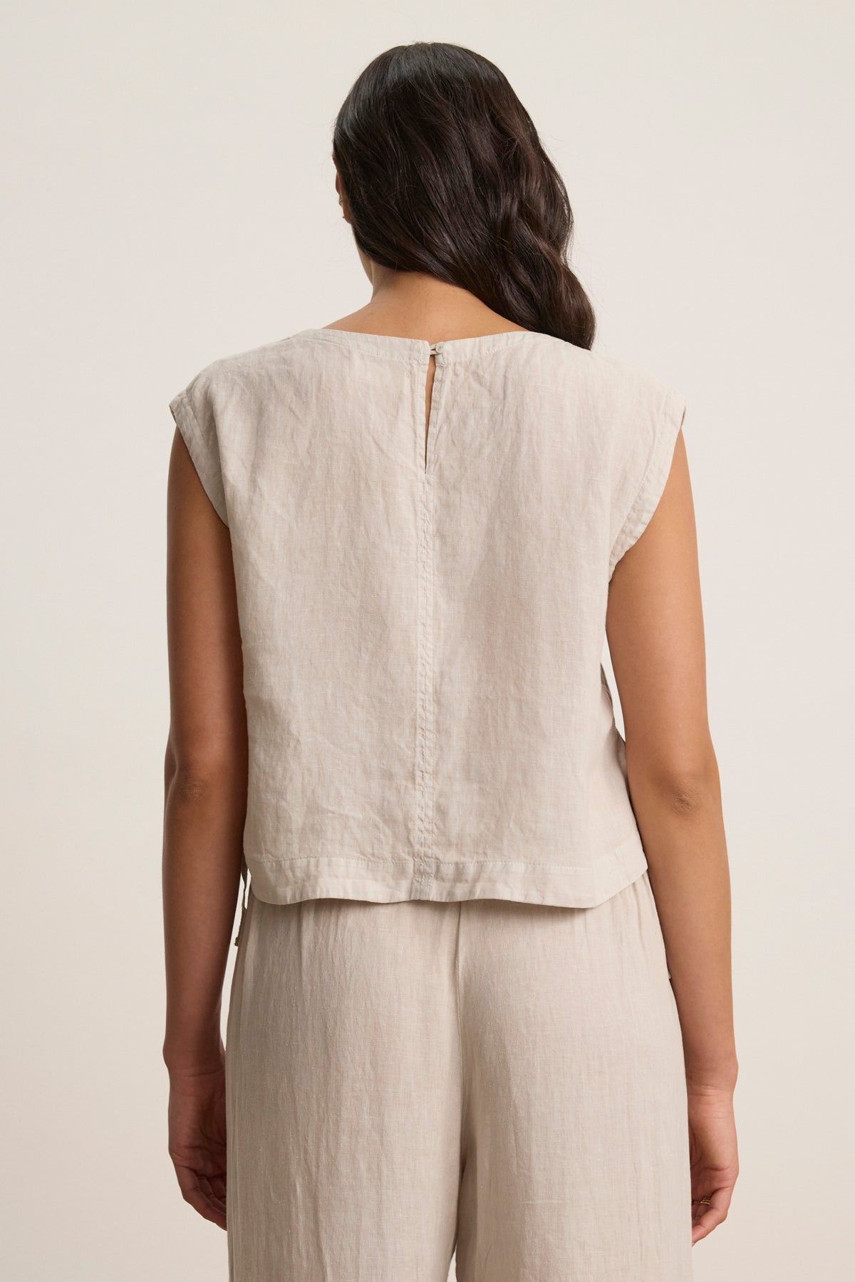 A person with wavy hair stands facing away, wearing the TAVIA LINEN TOP by Velvet by Graham & Spencer—a sleeveless, cropped light beige linen top—paired with matching pants.-39148767609025