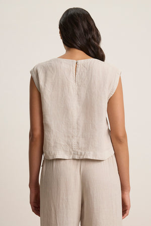 A person with wavy hair stands facing away, wearing the TAVIA LINEN TOP by Velvet by Graham & Spencer—a sleeveless, cropped light beige linen top—paired with matching pants.