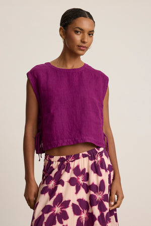 A woman wears the TAVIA LINEN TOP by Velvet by Graham & Spencer—a sleeveless purple linen piece—paired with a cream skirt with a purple floral print, highlighting its breezy feel and flowy silhouette against a plain backdrop.