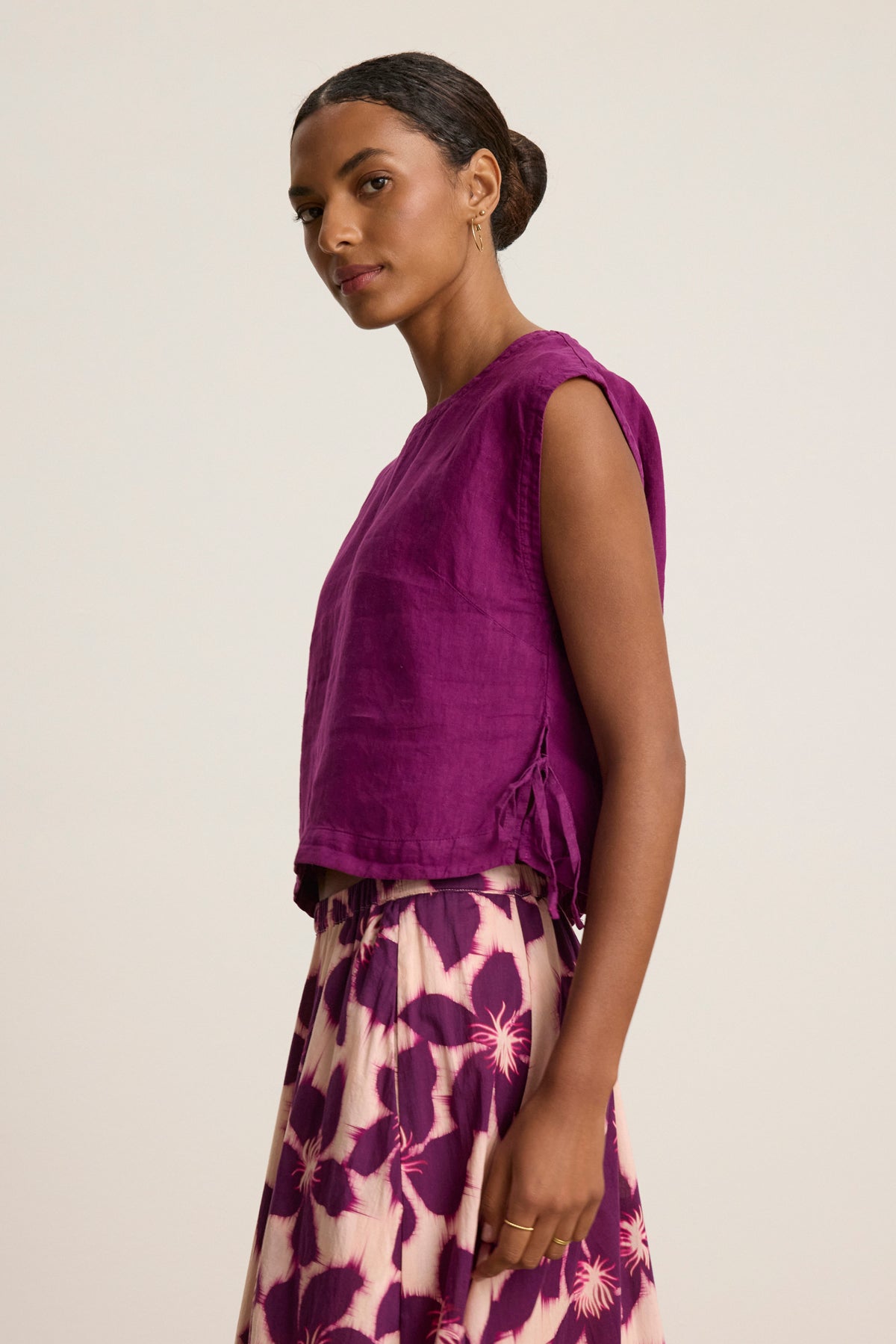 A woman stands in profile against a plain light background, wearing the sleeveless TAVIA LINEN TOP by Velvet by Graham & Spencer and a cream skirt with bold purple floral patterns.-39111855898817