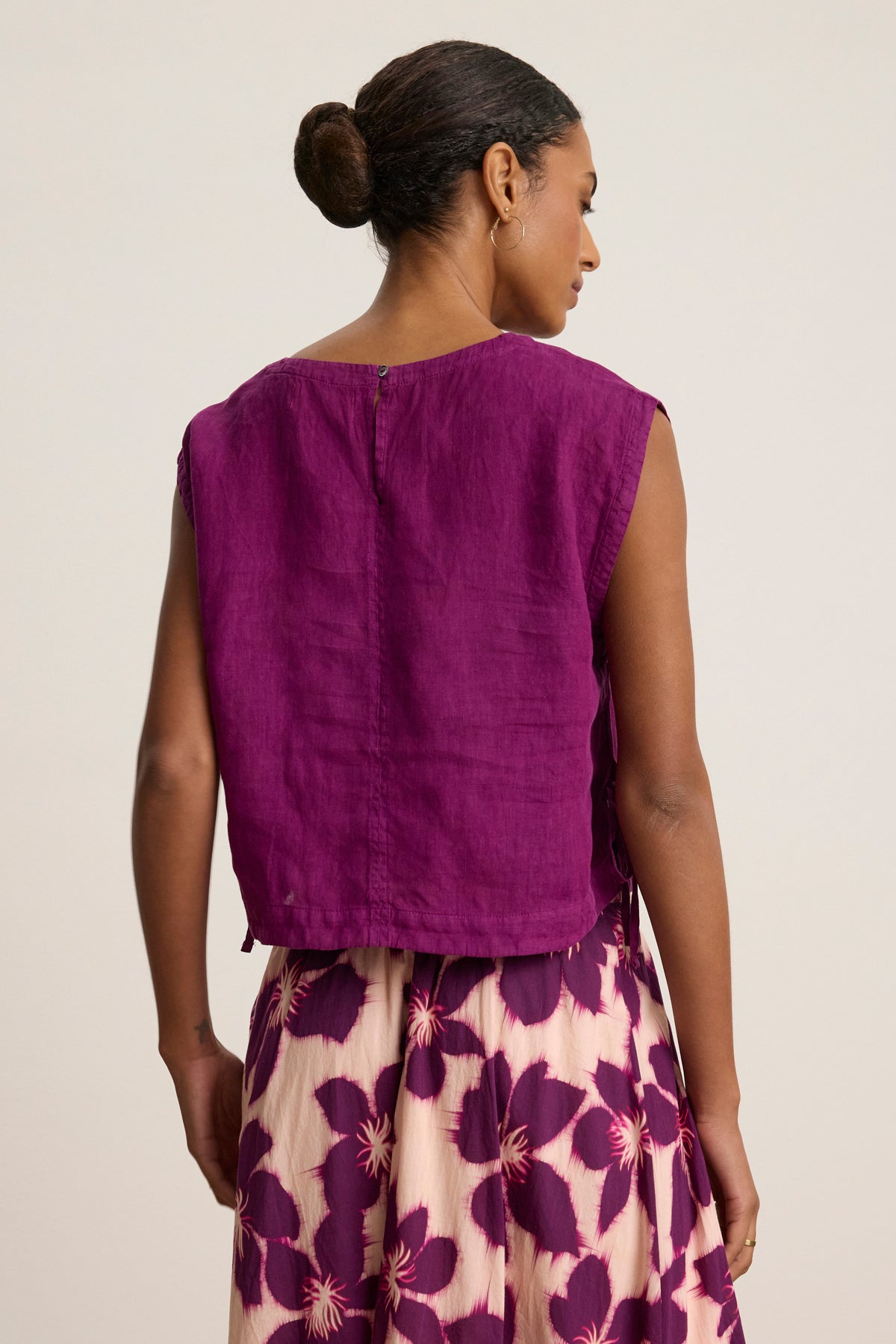 A woman with her hair in a bun models the TAVIA LINEN TOP by Velvet by Graham & Spencer, paired with a cream skirt featuring purple floral prints, shown from the back against a plain background.-39111856292033