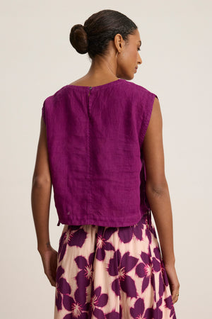 A woman with her hair in a bun models the TAVIA LINEN TOP by Velvet by Graham & Spencer, paired with a cream skirt featuring purple floral prints, shown from the back against a plain background.