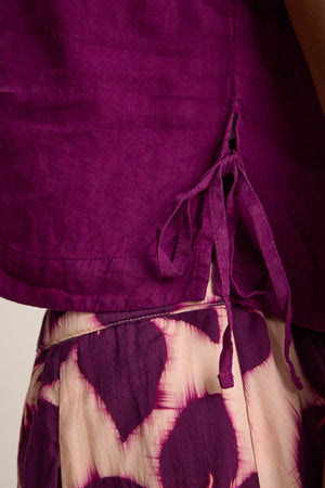 Close-up of a person wearing the TAVIA LINEN TOP by Velvet by Graham & Spencer, featuring side ties and paired with cream and purple patterned pants for a flowy silhouette.