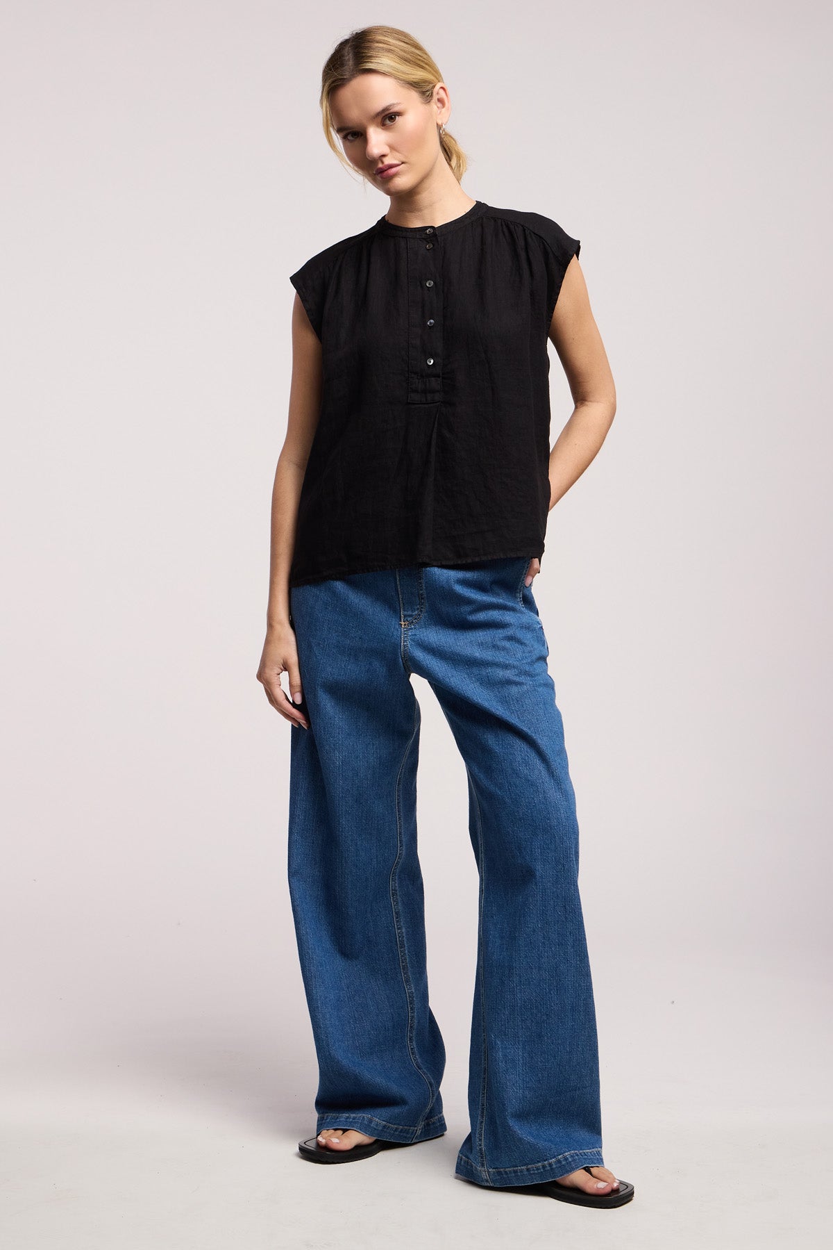 A person stands against a plain background wearing the BEC LINEN TOP by Velvet by Graham & Spencer, styled with wide-leg blue jeans and black sandals.-41526929227969