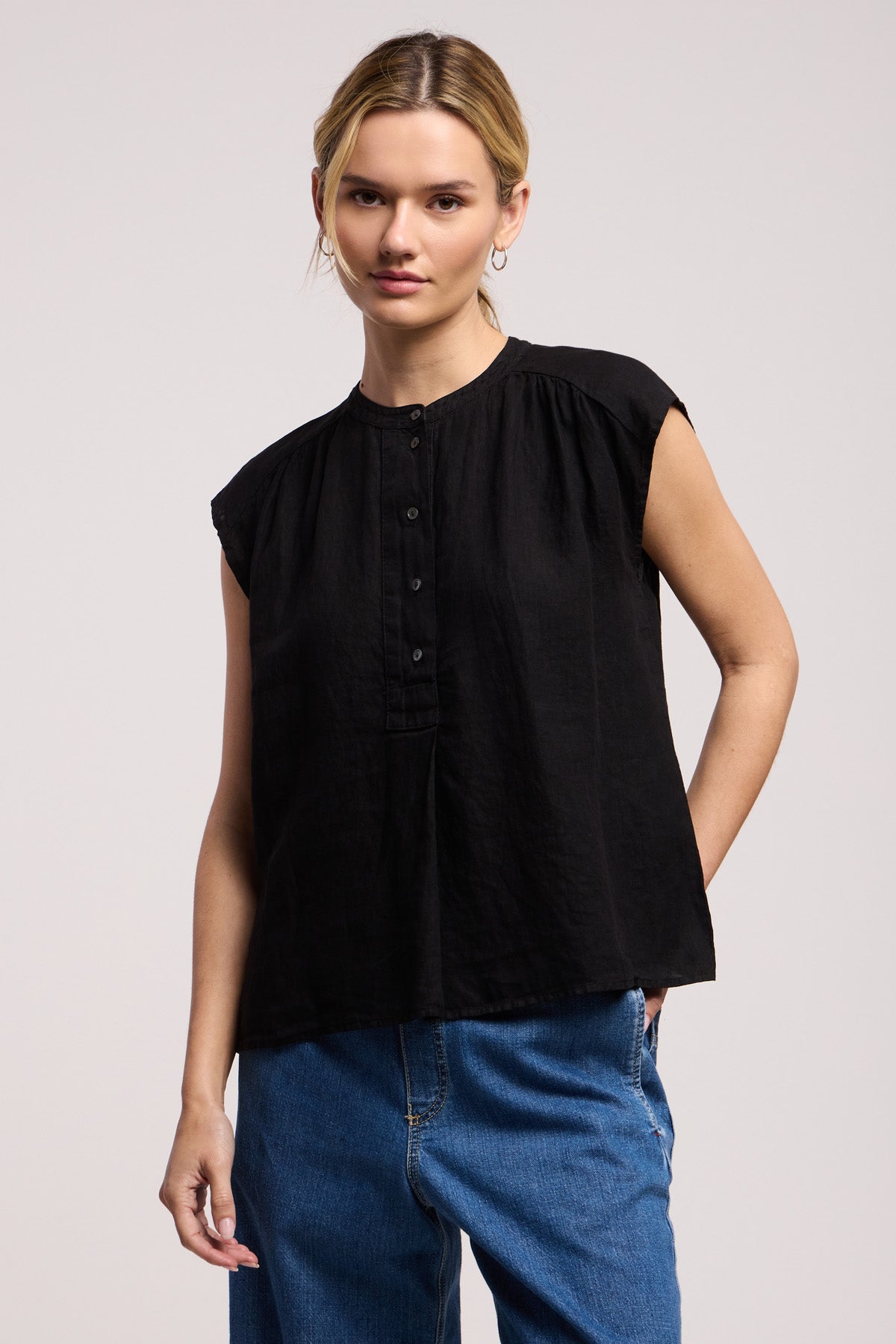 Someone wearing the Velvet by Graham & Spencer BEC LINEN TOP in black with blue jeans stands against a plain light background.