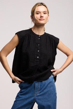 A person wearing the BEC LINEN TOP by Velvet by Graham & Spencer in black with blue jeans stands with one hand in a pocket against a plain background.