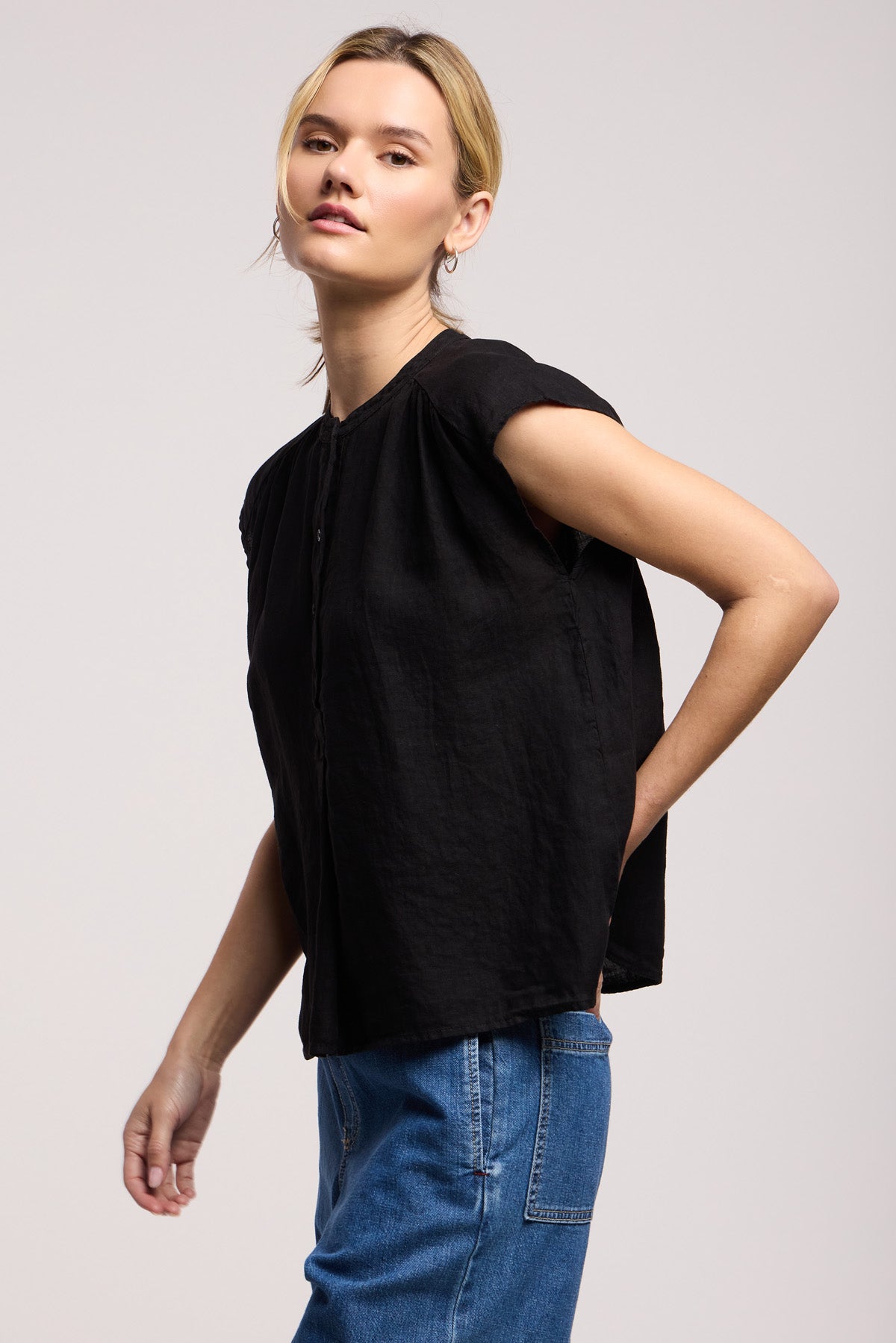 Someone wearing the Velvet by Graham & Spencer BEC LINEN TOP in black with blue jeans stands against a plain light background, one hand behind their back.