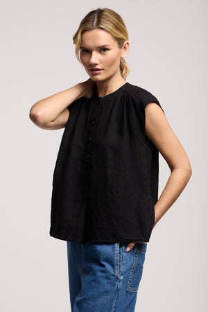 A person models the BEC LINEN TOP by Velvet by Graham & Spencer, a loose-fitting black sleeveless linen top, paired with blue jeans against a plain background, one hand in their pocket and the other touching their neck.