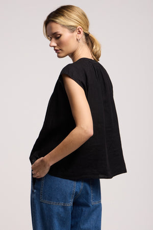 A person stands sideways, wearing the BEC LINEN TOP by Velvet by Graham & Spencer—a loose black sleeveless linen top styled with blue jeans. The boxy silhouette and plain backdrop highlight this modern, effortless summer look. Hair is tied back, gaze downward.
