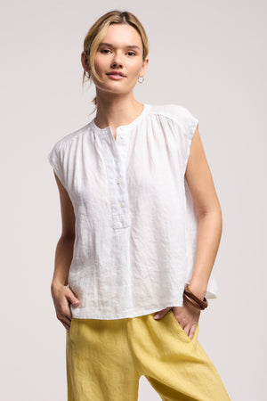 A model wears the BEC LINEN TOP by Velvet by Graham & Spencer, paired with yellow pants and posing with one hand in their pocket against a plain background.
