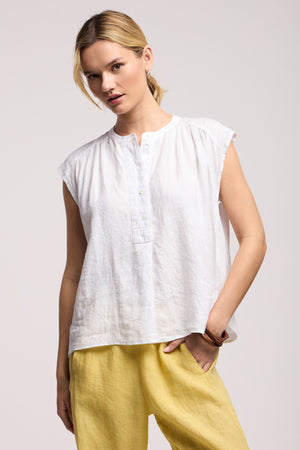 A person models the Velvet by Graham & Spencer BEC LINEN TOP, a white sleeveless summer piece, paired with light yellow pants, standing with one hand in their pocket against a plain background.